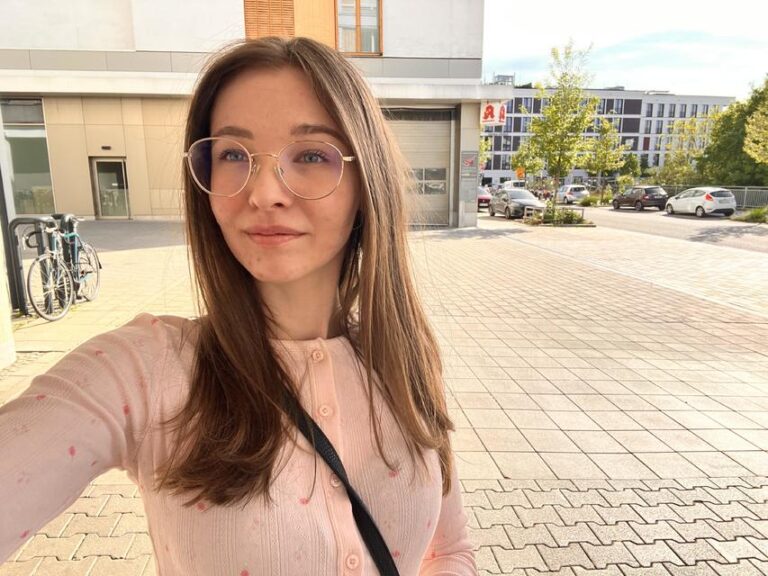 Emmi Hill macht ein Selfie im Sonnenschein auf einem Hof, trägt Brille und ein schickes Outfit.