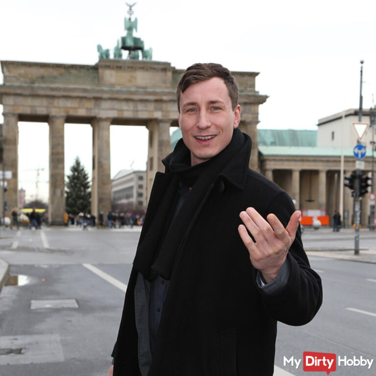 Jason Steel Erotikdarsteller steht mit einladender Geste vor dem Brandenburger Tor in Berlin.