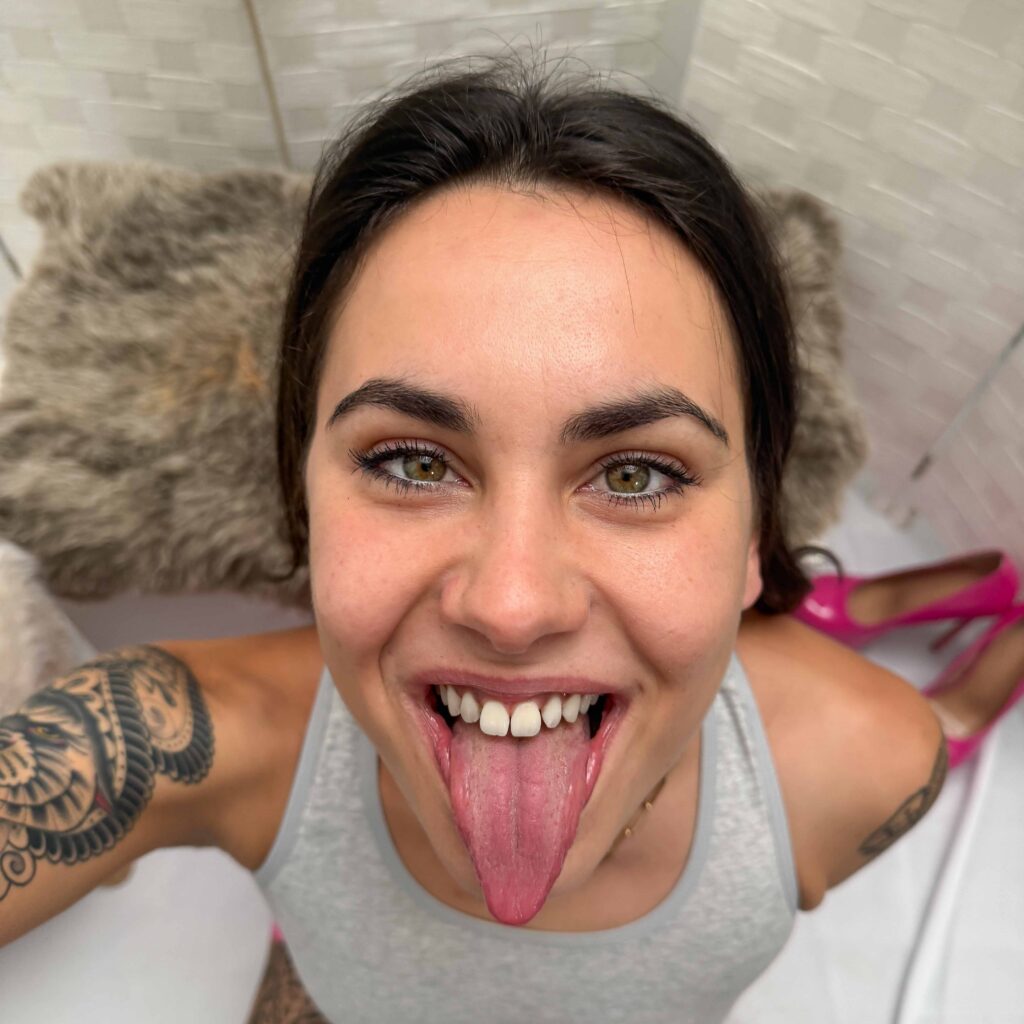 Jenny Stella in weißem Tanktop, mit verspieltem Gesichtsausdruck – schielender Blick und herausgestreckte Zunge in einer charakteristischen Pose. Deutsche Pornodarstellerin