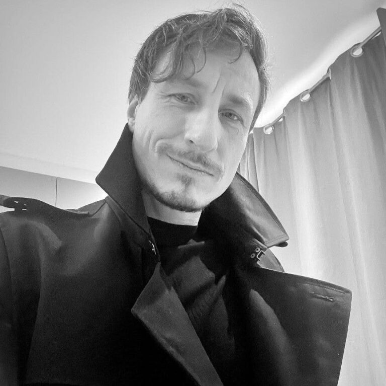 Jason Steel in Schwarz-Weiß mit hochgeschlagenem Mantelkragen und ernstem Blick in die Kamera