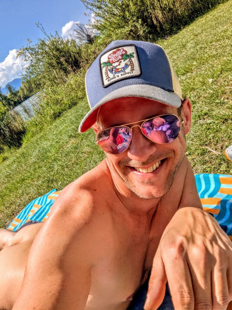Larry Ragnarök Adler entspannt lachend nackt am FKK-Strand in Happing mit Sonnenbrille und Cap – dein erfahrener Sexperte für Lust und weibliche Ejakulation und ist selbst eine Fremdsprachen-Queen