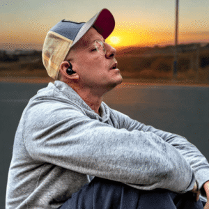 Larry Adler sitzt am Straßenrand vor Sonnenuntergang, reflektiert seine Vision und Ängste, umgeben von Natur, mit Cap, grauem Hoodie und blauer Jogginghose.